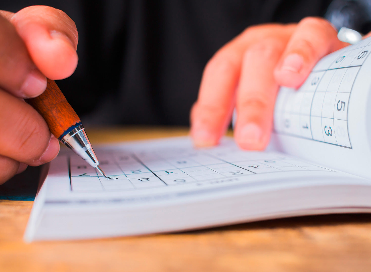 Nahaufnahme von Händen, die mit einem Bleistift ein Sudoku-Rätsel in einem Heft ausfüllen