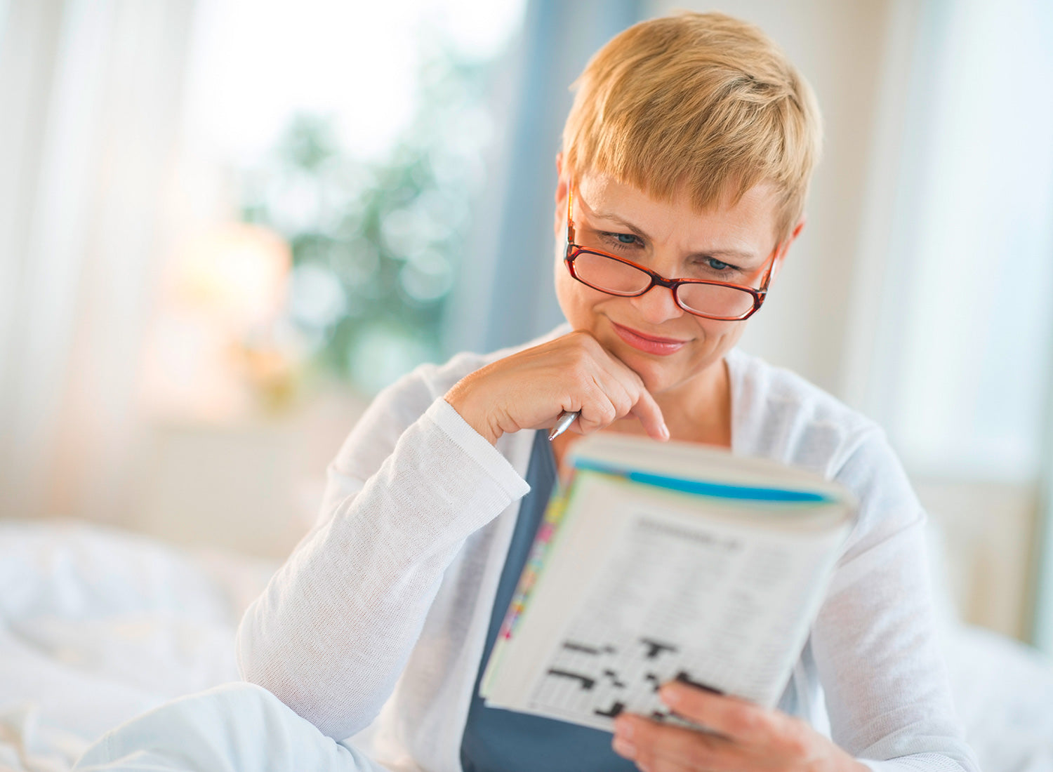 Person mit kurzem blondem Haar sitzt im Bett und löst Kreuzworträtsel in einem Heft